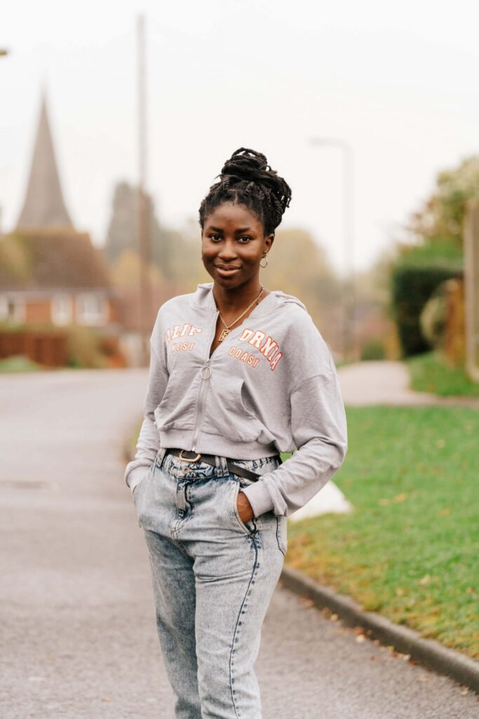 Young woman smiling at camera wearing gray zip-up hoodie and jeans, standing on street with blurred buildings and church spires in background.