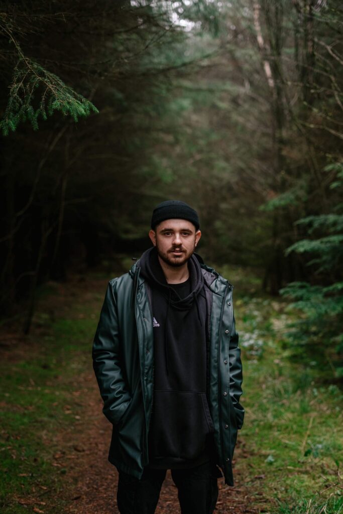 Man in black beanie and dark jacket standing on forest path surrounded by tall evergreen trees.